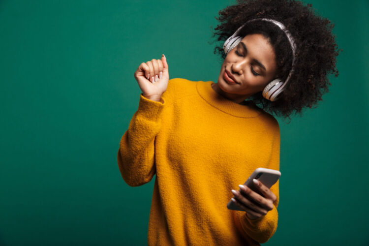 Image of a beautiful african curly woman posing isolated over dark blue wall background listening music with headphones using smartphone.