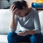distressed young woman on couch at home looking down at phone - substance use and mental health disorders, COVID-19