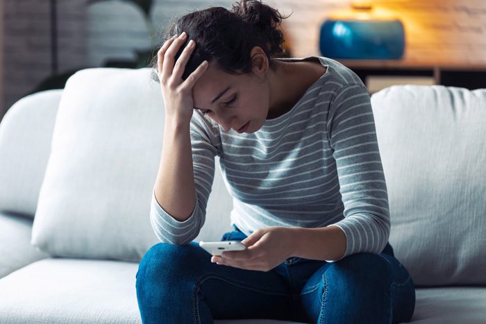 distressed young woman on couch at home looking down at phone - substance use and mental health disorders, COVID-19